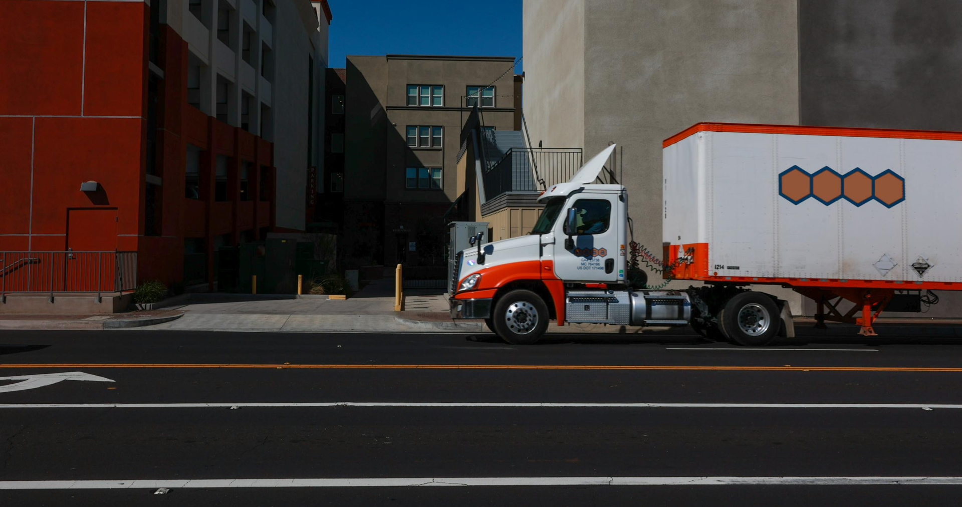 A delivery truck parked on a city street under bright sunlight in front of modern buildings.